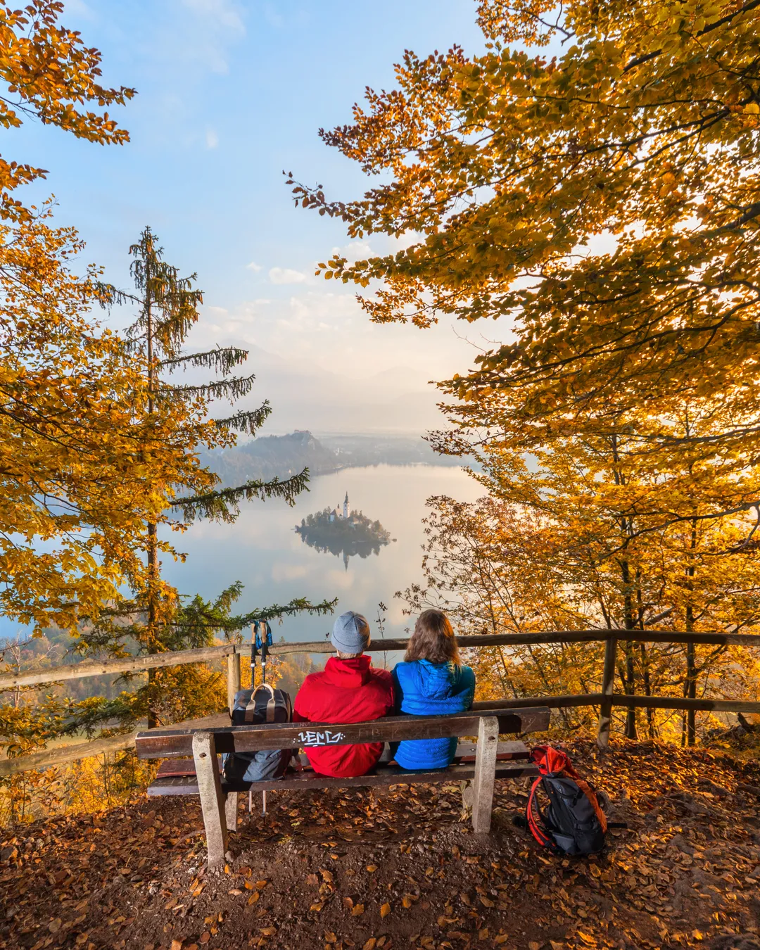 Two walkers on a bench above Lake Bled, framed by autumn beeches