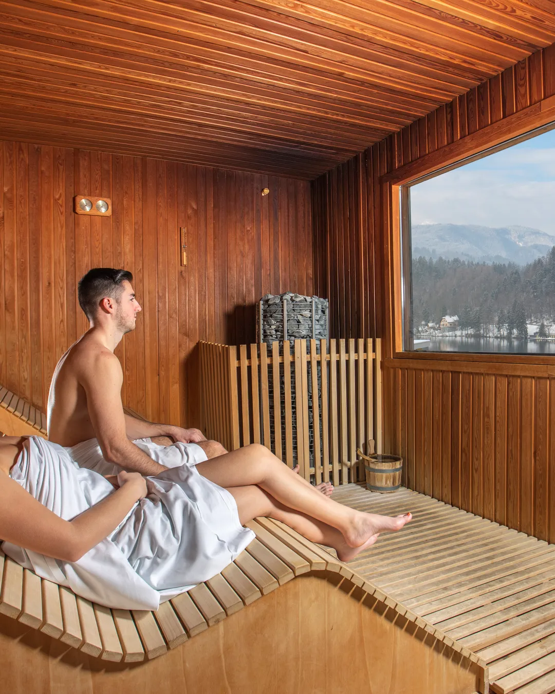 Two guests in a wooden sauna, the window looking out over Lake Bled
