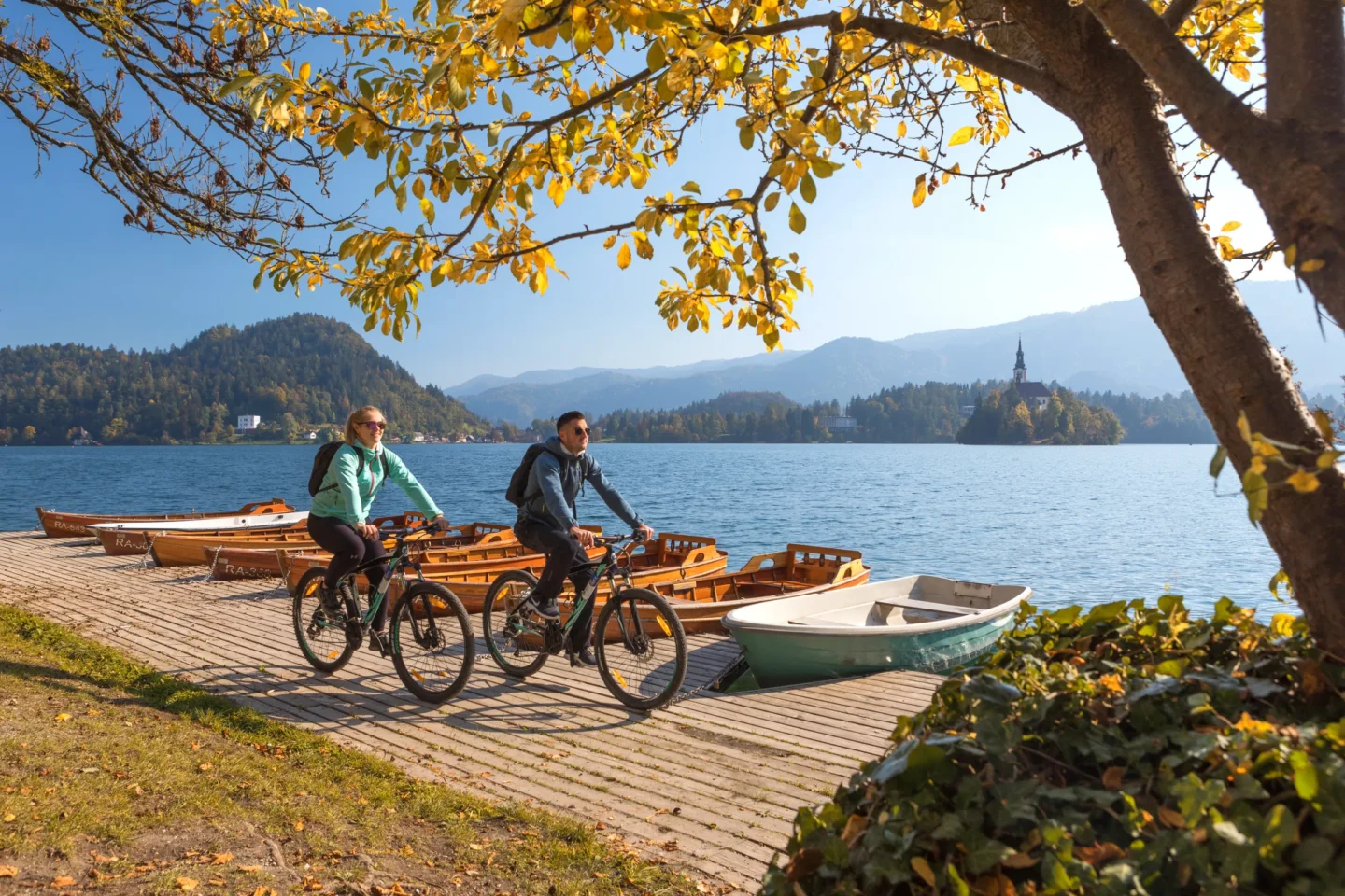 Two cyclists on a wooden jetty on Lake Bled in autumn, rowing boats moored alongside, the island church behind