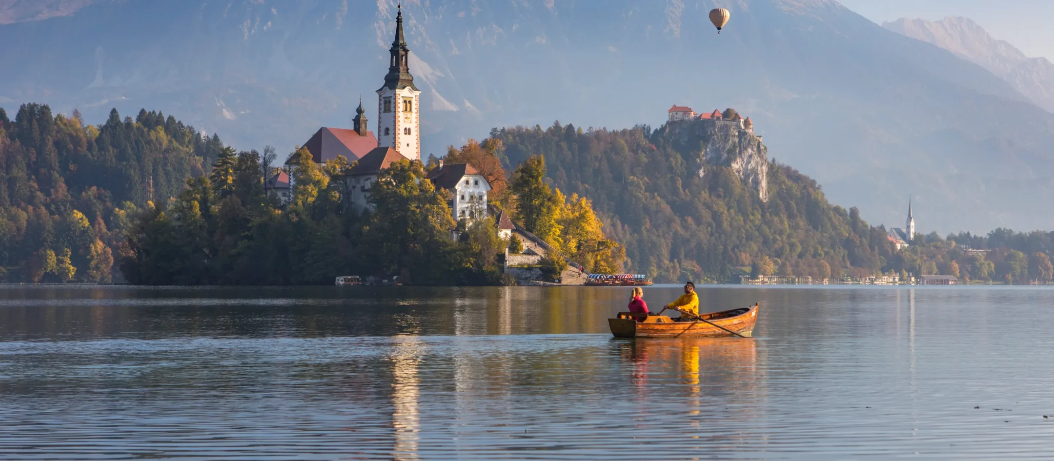 Two people rowing a wooden boat on Lake Bled at sunrise, the island church and castle behind